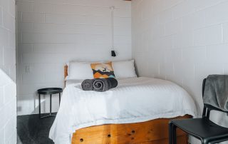 A double bed pictured with crips white linen, a colourful show pillow and two rolled towels. There is a bedside table and a chair pushed up against the wall.