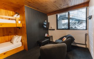 A room featuring wooden bunk beds, a seating area with chairs, a TV mounted on the wall, and a large window with a view of the snowy outdoors.