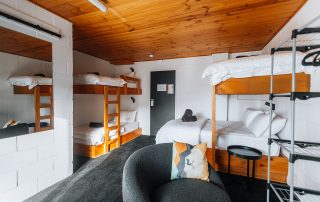 A room featuring multiple wooden bunk beds, a cushioned chair with a decorative pillow, and a mirrored wall reflecting the room’s layout.