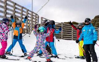 A group of children in colorful ski outfits standing on a snowy slope with their ski poles raised, accompanied by an instructor in a blue jacket.