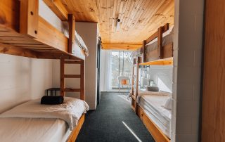 Two sets of bunk beds in the foreground looking out to a sliding door with a balcony. There is a chair pictured in front of the window that looks out to a picturesque snowy mountains bushland.