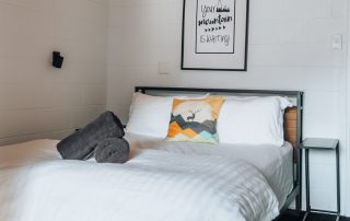 A cozy queen bed with white linens, a decorative pillow, and folded towels placed on top in a room with a wooden ceiling and white walls.