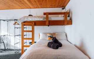 A bunk bed featuring a double on the bottom with a show pillow and two rolled towels. There is a single bed on top with white linen and towels, as well as a storage rack to the left of the beds.
