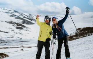 Two skiers in winter gear taking a selfie on a snowy mountain slope with a scenic backdrop of snow-covered hills and a blue sky.